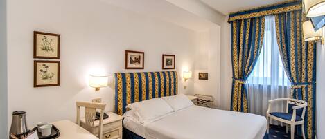 Une chambre d’hôtel comprenant un lit, deux tables de chevet, un bureau avec une chaise, une lampe et une fenêtre avec des rideaux.