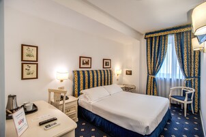 A hotel room with a bed, two bedside tables, a desk with a chair, a lamp, and a window with curtains.