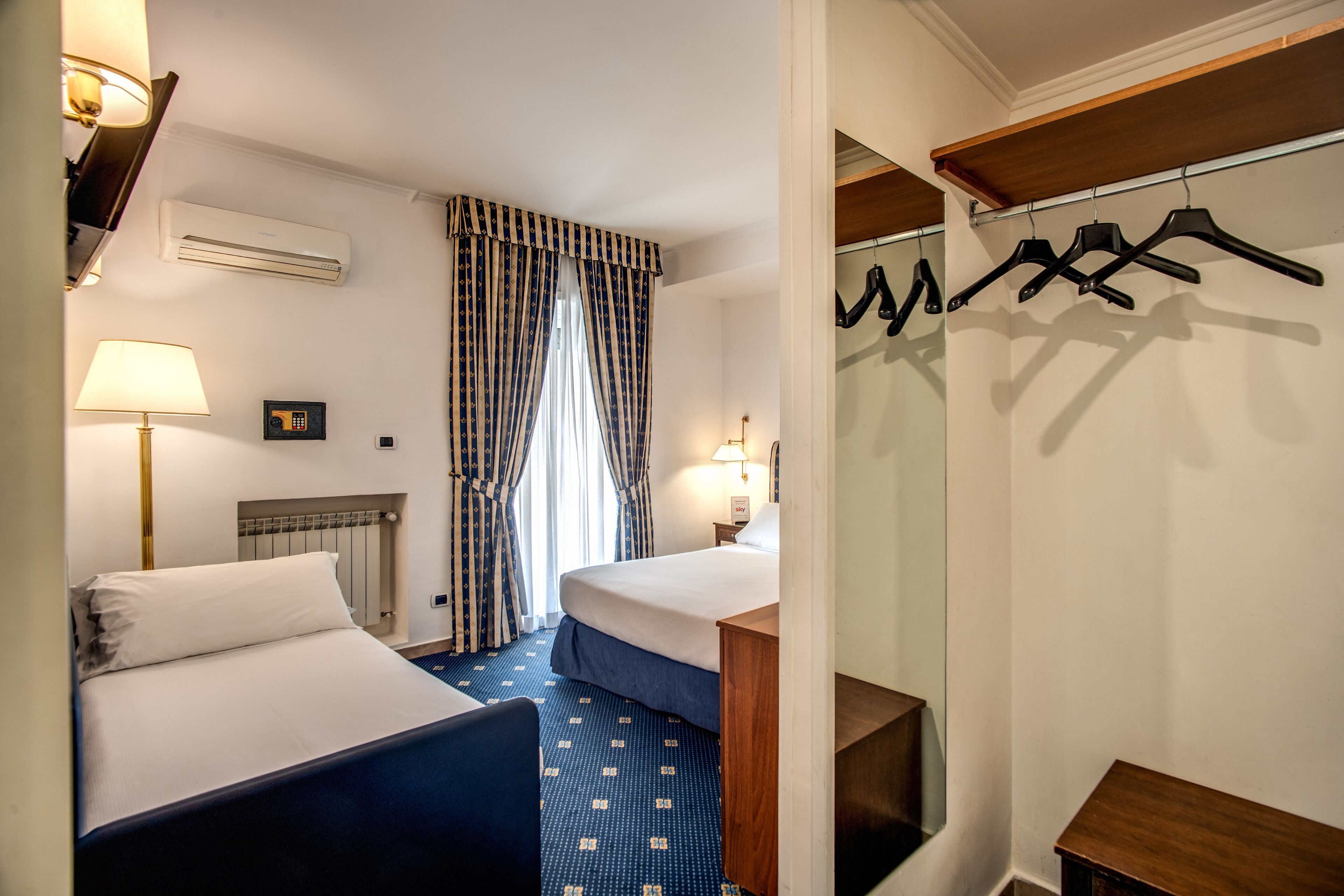 A hotel room with two beds, a desk, a chair, a lamp, an air conditioner, a radiator, a clock, and a coat rack.
