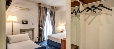 A hotel room with two beds, a desk, a chair, a lamp, an air conditioner, a radiator, a clock, and a coat rack.