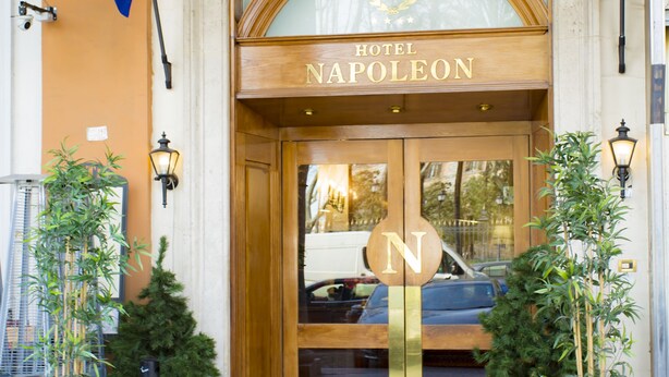 La entrada del Hotel Napoleon, con una gran puerta de madera, un ventanal encima y un cartel con el nombre del hotel. Hay dos macetas con plantas a cada lado de la puerta.