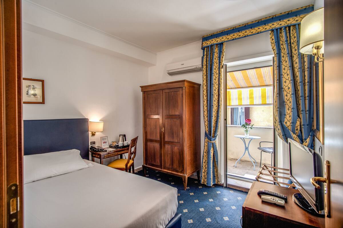 Una habitación de hotel con una cama, un escritorio con lámpara, un armario, una mesita con un florero y un ventanal con cortinas.