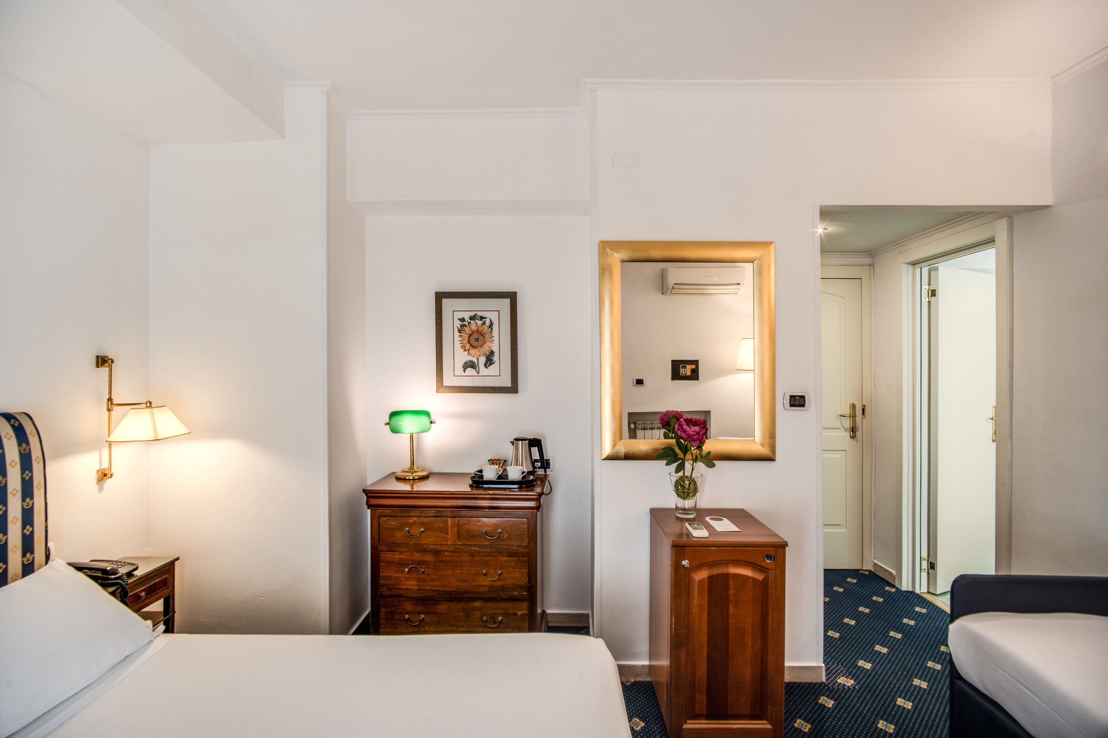 A hotel room with a bed, a desk with a lamp, a dresser, a mirror, and a door leading to another room.