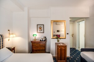 A hotel room with a bed, a desk with a lamp, a dresser, a mirror, and a door leading to another room.