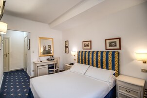 A hotel room with a bed, a desk, a chair, a nightstand, and a wall-mounted lamp.