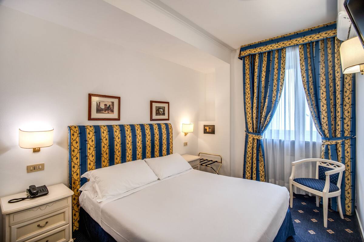 Una habitación de hotel con cama, mesitas de noche, una silla y cortinas.