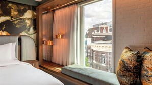 A hotel room with a large window, a bed, a bench, and a view of buildings outside.