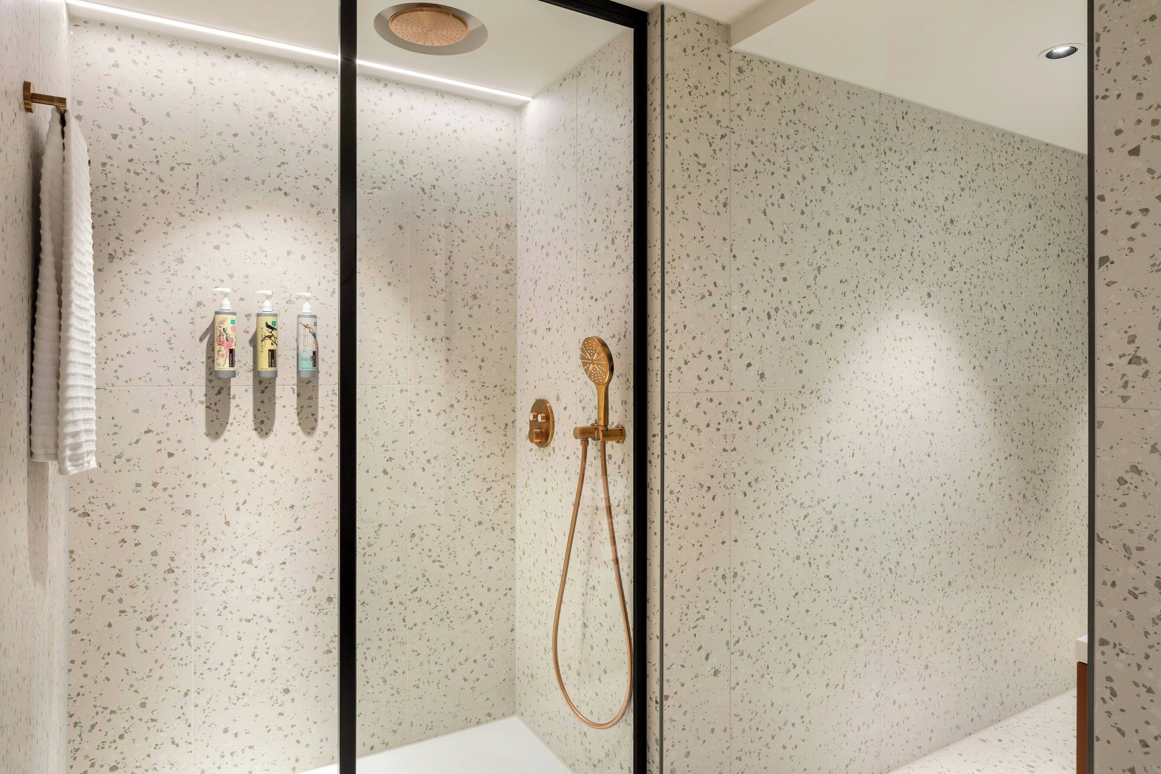 A modern bathroom with a speckled tile wall, a glass shower enclosure, and a white bathtub.