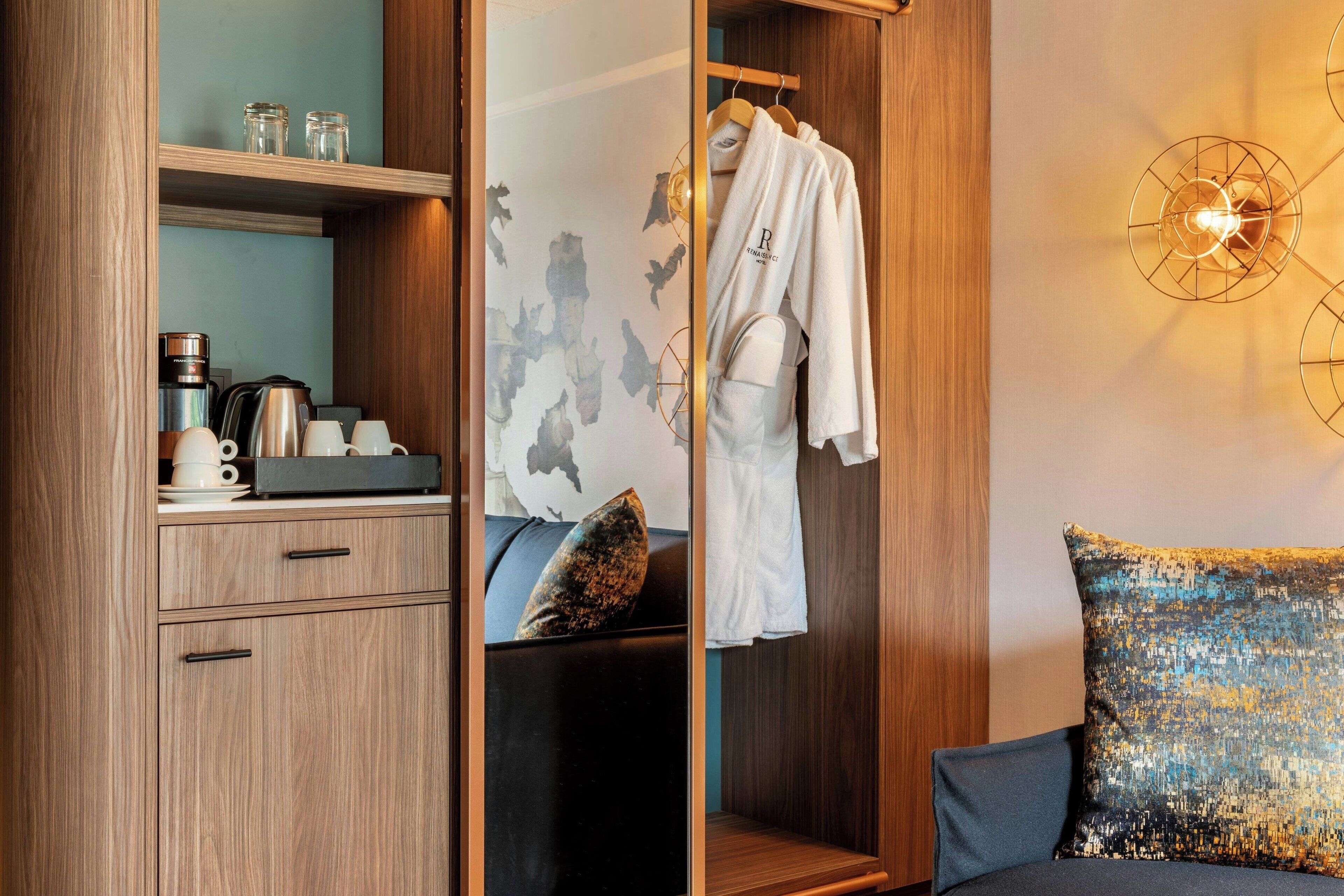 Habitación de hotel con un armario de madera, una cama con almohada azul, una bata blanca colgada en un perchero y una lámpara de pared decorativa.
