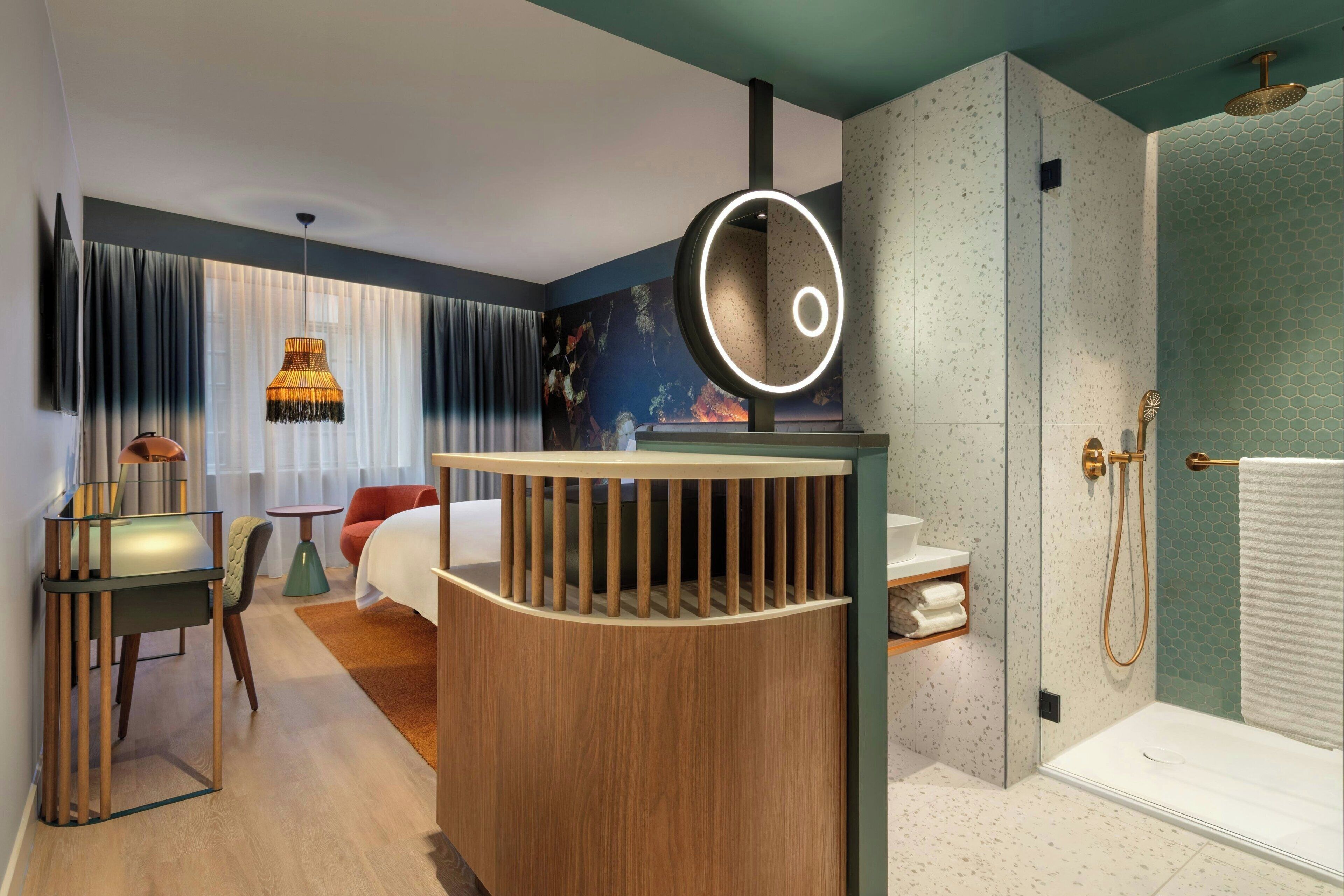 A modern hotel room with a curved wooden partition, a round mirror with a light, a shower with a gold showerhead, and a bed with a red chair.