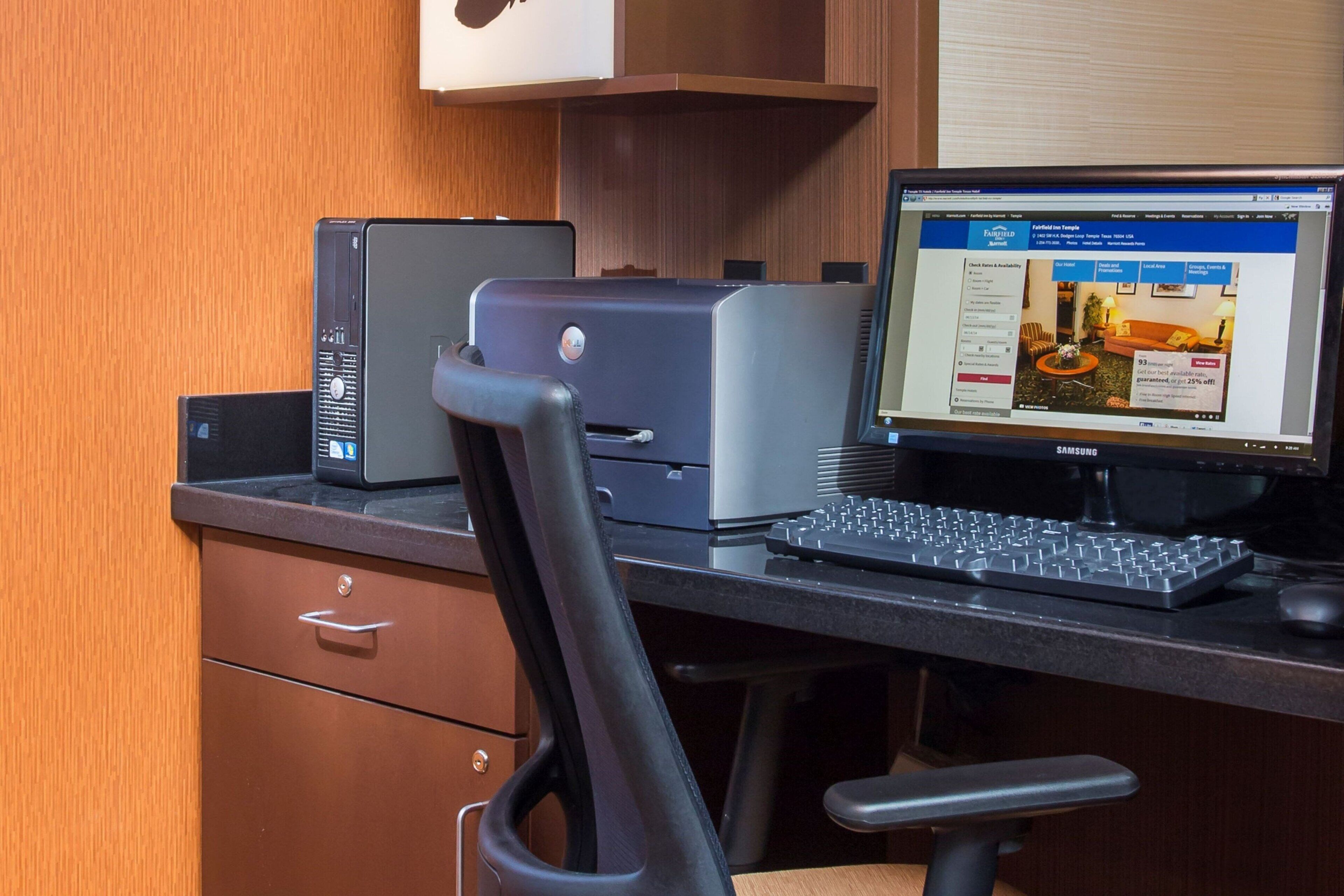 A hotel room with a desk, a desktop computer, a printer, and a chair.