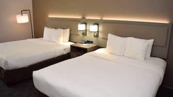 Habitación de hotel con dos camas, cada una con ropa blanca de cama y almohadas, una mesita de noche con lámpara y luces de lectura en la pared.