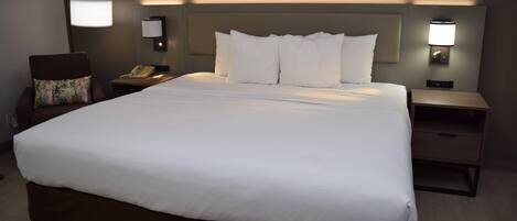 A large bed with white linens, two bedside tables with lamps, a chair with a floral pillow, and a wall-mounted phone.