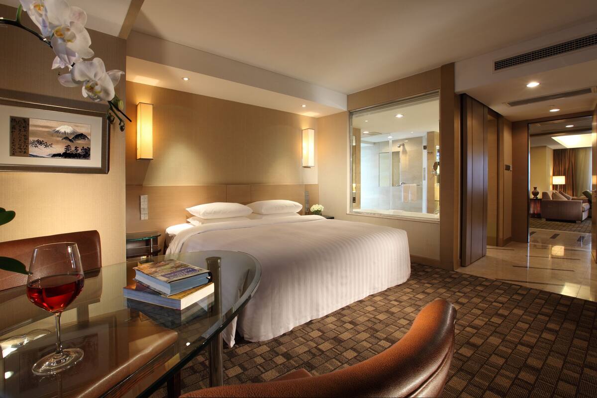A hotel room with a bed, bedside table, and a glass table with a wine glass and books.