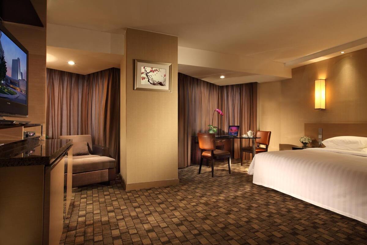 A hotel room with a bed, a desk with a computer, a chair, a television, and a wall-mounted lamp.