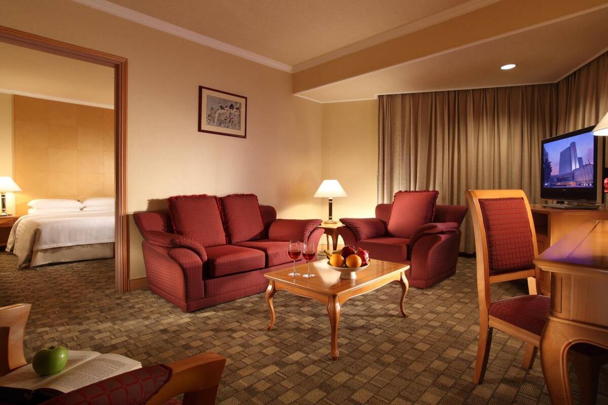 A hotel room with a red sofa, a wooden coffee table with fruit and wine glasses, a TV, and a bed with white linens.