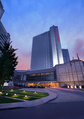 A modern hotel building with a prominent vertical sign, a well-maintained garden area, and a clear sky.