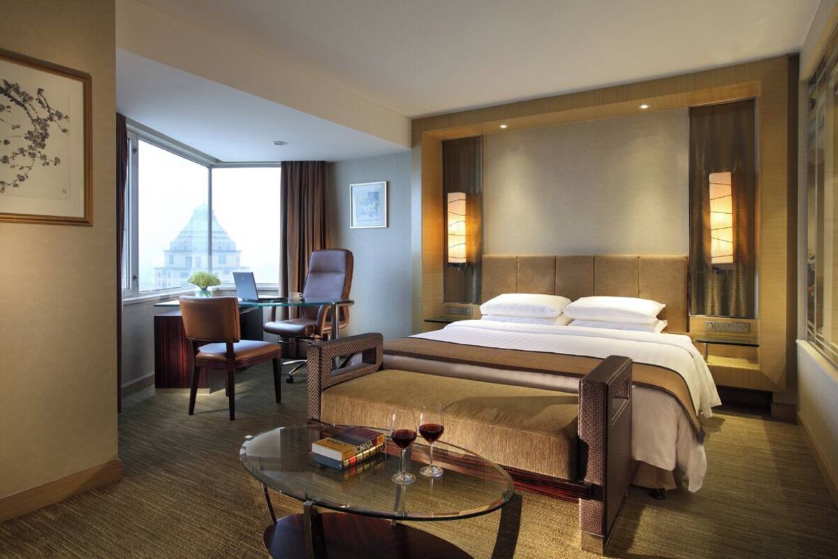 A hotel room with a large bed, a desk with a chair, a glass table with two wine glasses and a book, and a view of a building through a large window.