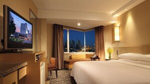 A hotel room with a large bed, a TV on a wooden dresser, a sofa, a small table with a vase, and a window with a city view. - Hotel New Century Beijing (Beijing)