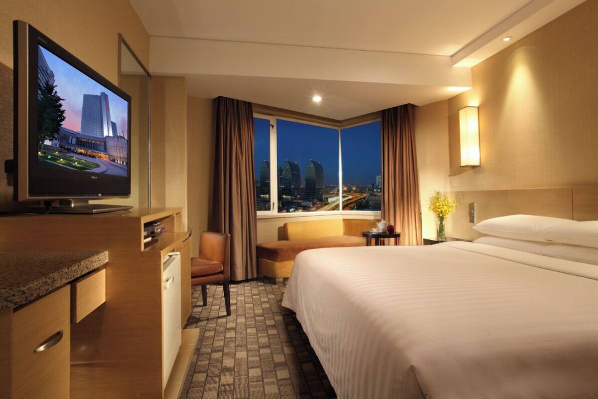 A hotel room with a large bed, a TV on a wooden dresser, a sofa, a small table with a vase, and a window with a city view.