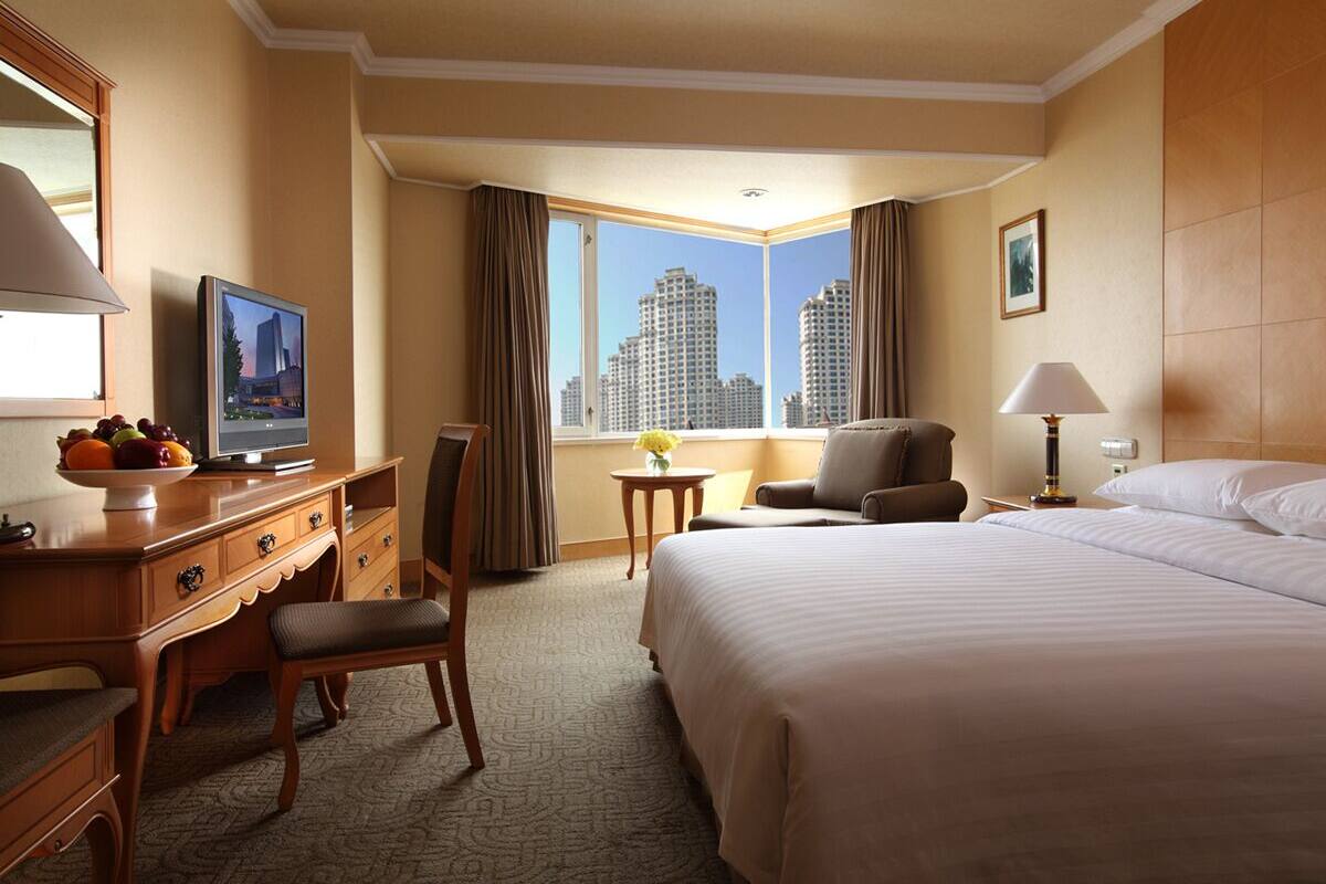 A hotel room with a large bed, a desk with a television, a chair, a small table with a vase, and a view of tall buildings.