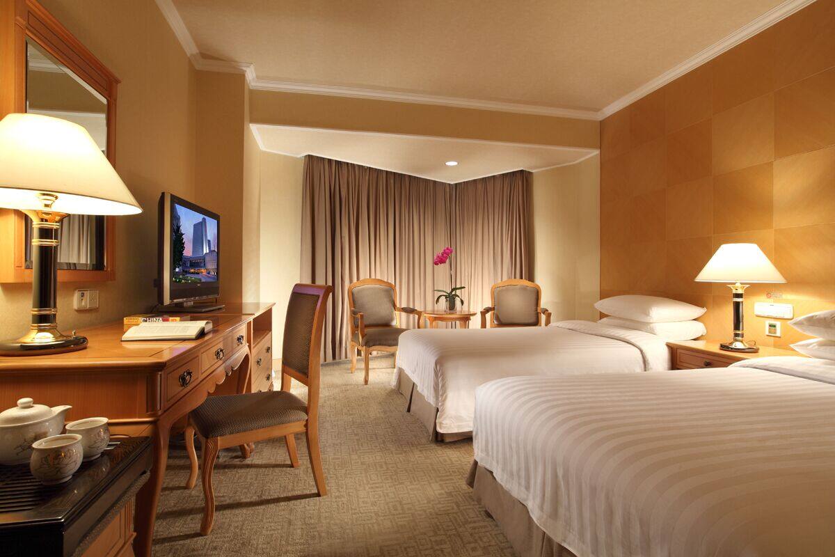 A hotel room with two beds, a desk with a TV, a chair, a nightstand with a lamp, and a large window with curtains.