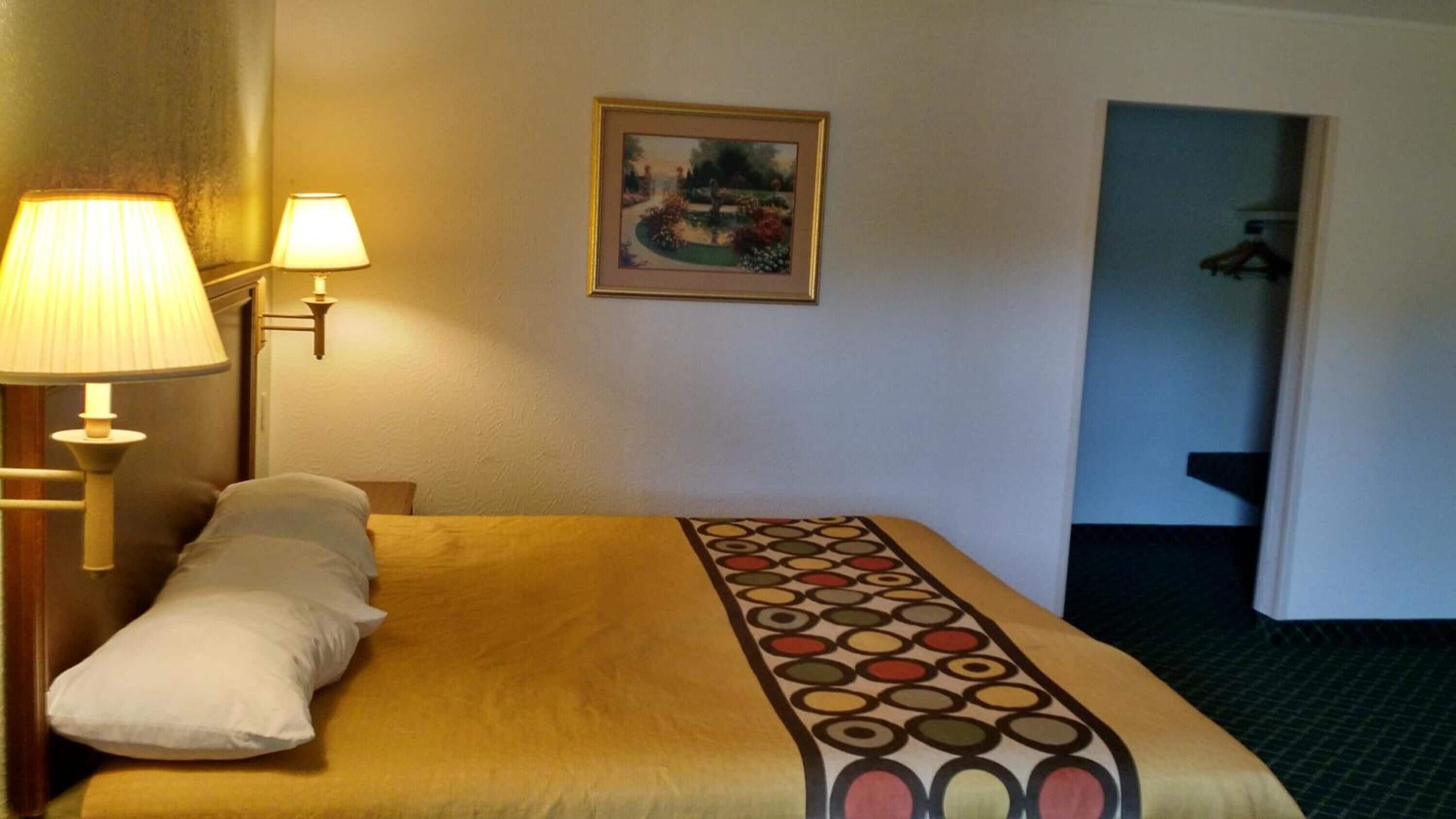 A hotel room with a bed, two bedside lamps, a framed picture on the wall, and a door leading to another room.