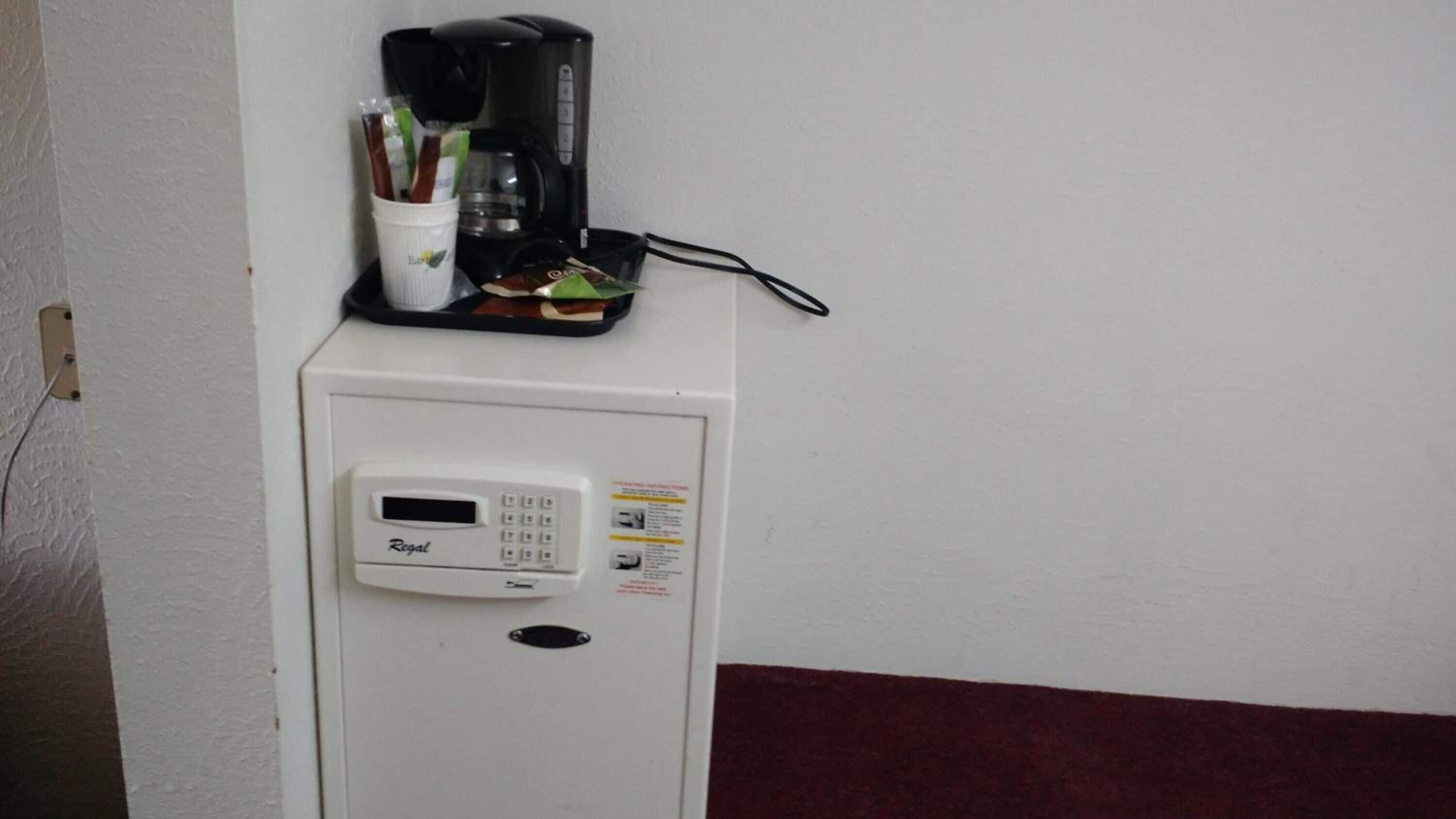 Un refrigerador blanco con una cafetera encima, una taza pequeña con utensilios y un cable de alimentación conectado.