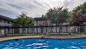 A swimming pool with a metal ladder, surrounded by a white fence, and a building with balconies in the background.