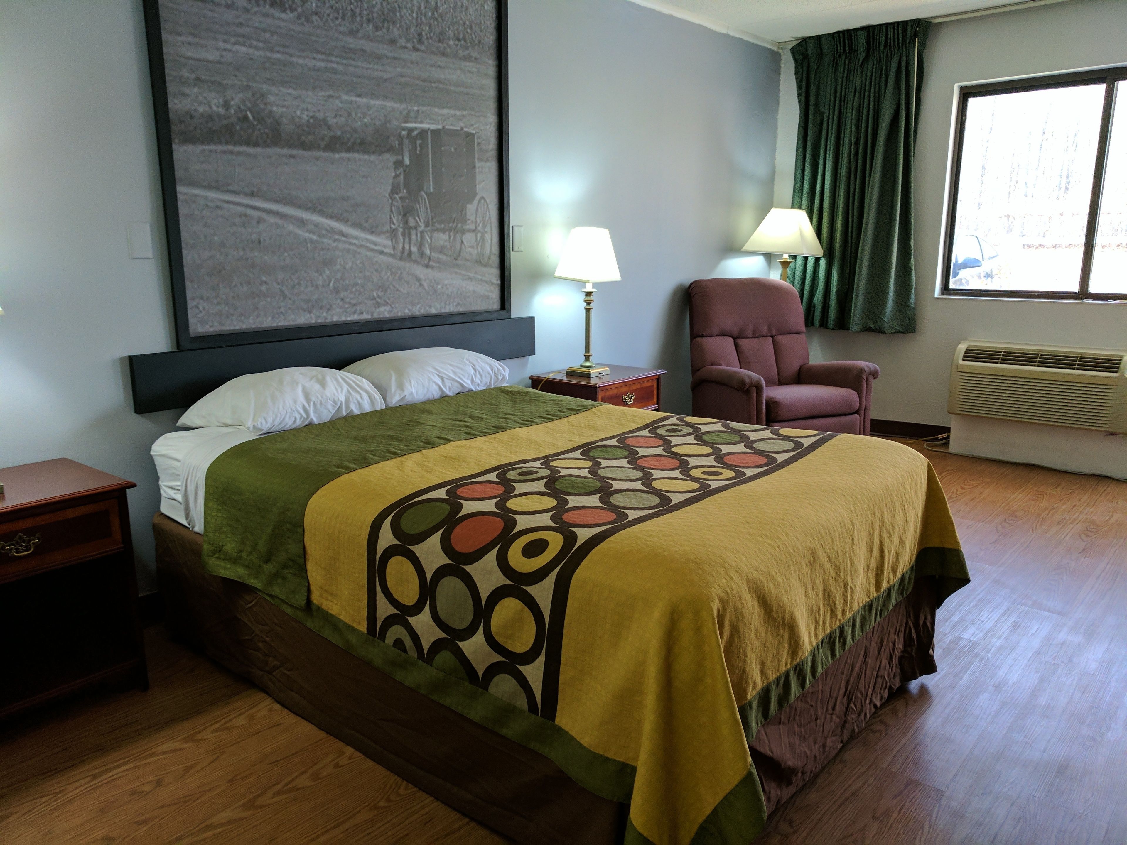 Habitación de hotel con cama, una silla, mesita de noche, lámpara y un cuadro grande en la pared.