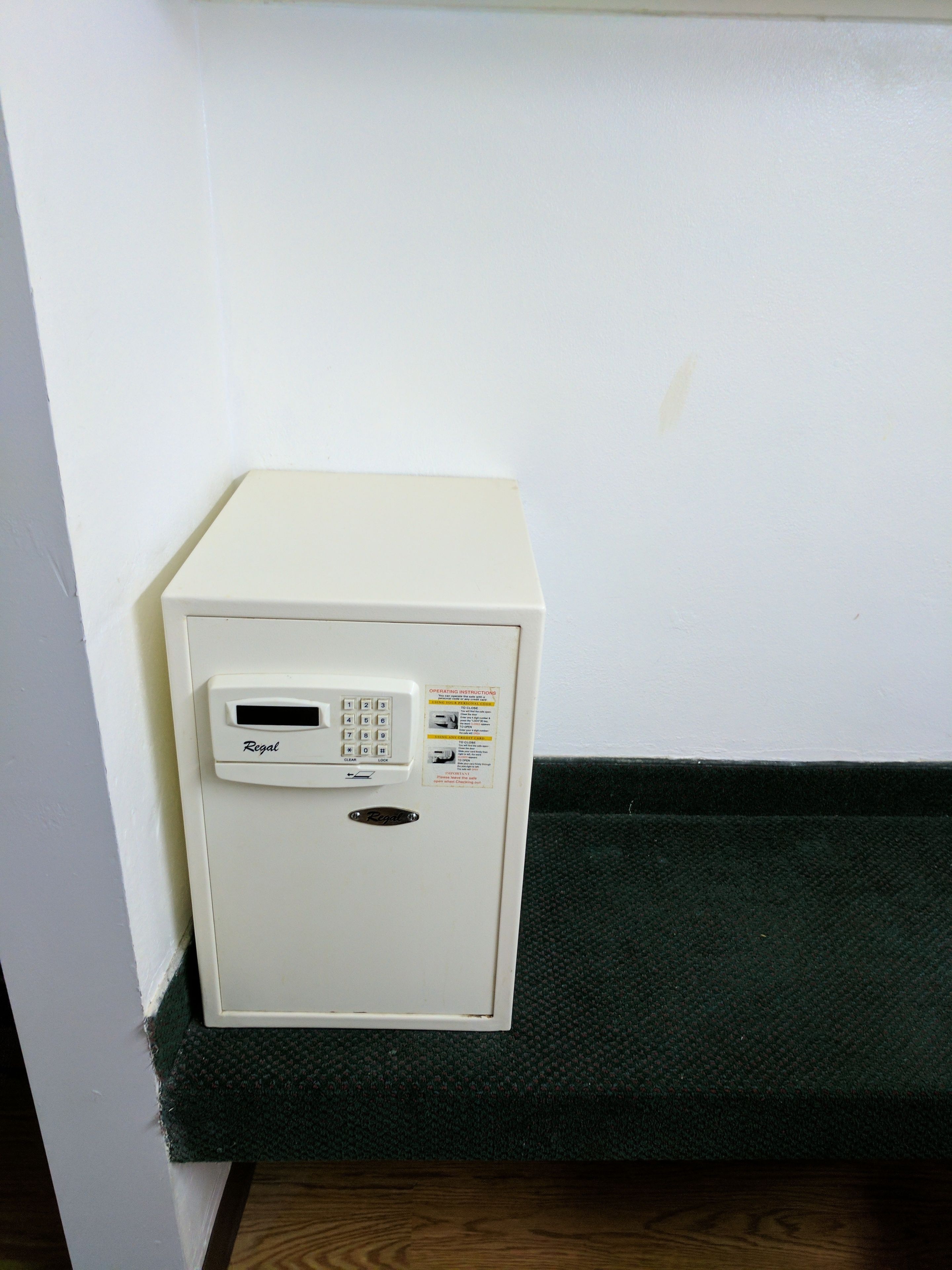 A white minibar with a digital keypad and a small screen, placed against a white wall with a green textured area below.