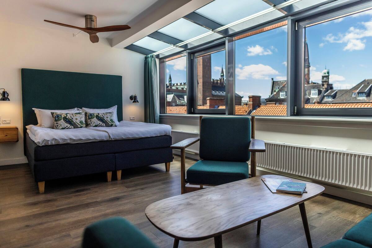 A modern bedroom with a bed, a chair, a table, and a view of the cityscape through large windows.