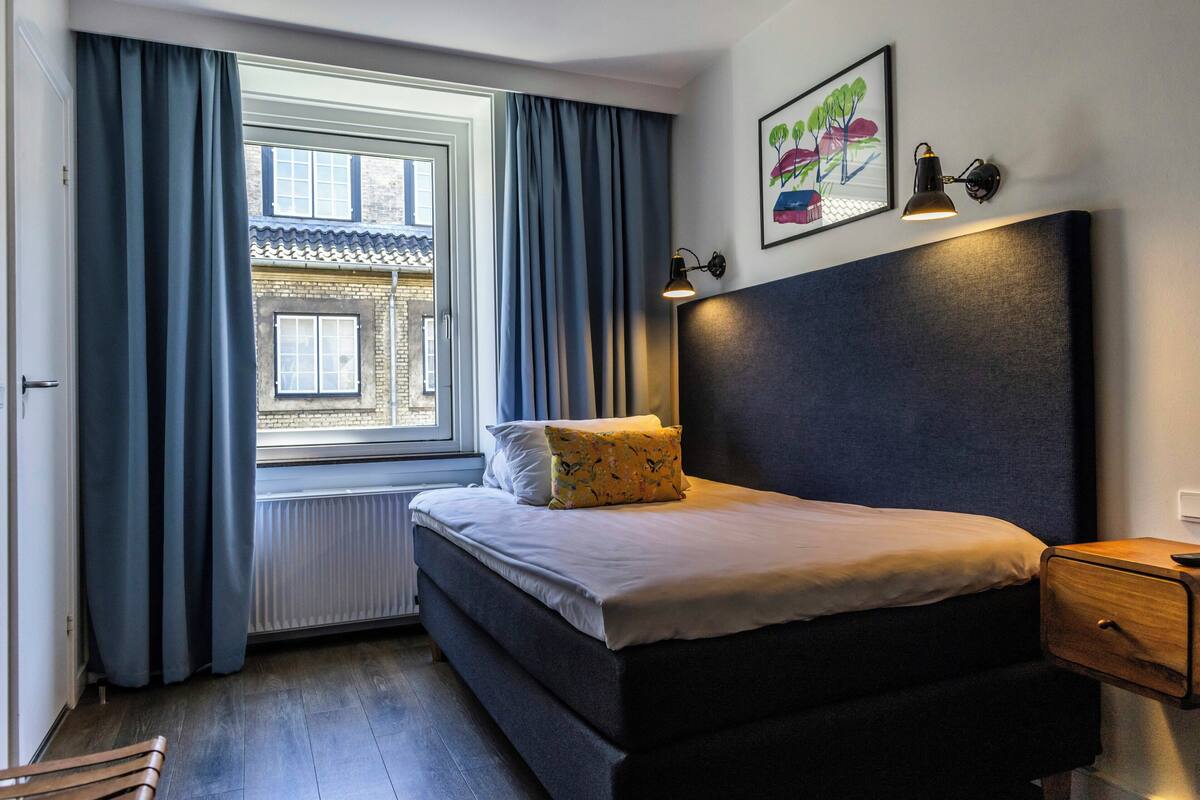 A bedroom with a bed, a nightstand, a window with curtains, a radiator, and a wall-mounted lamp.