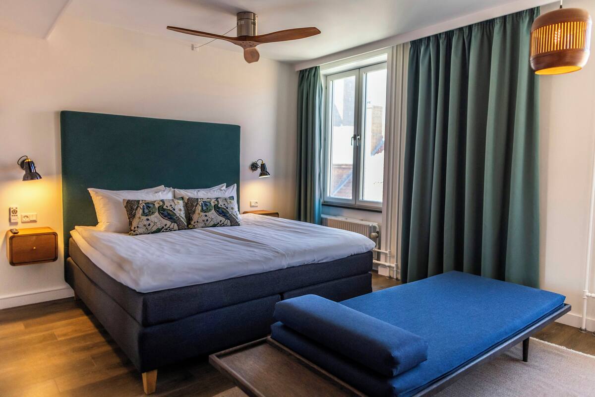 A bedroom with a bed, bedside lamps, a ceiling fan, a window with curtains, and a bench.