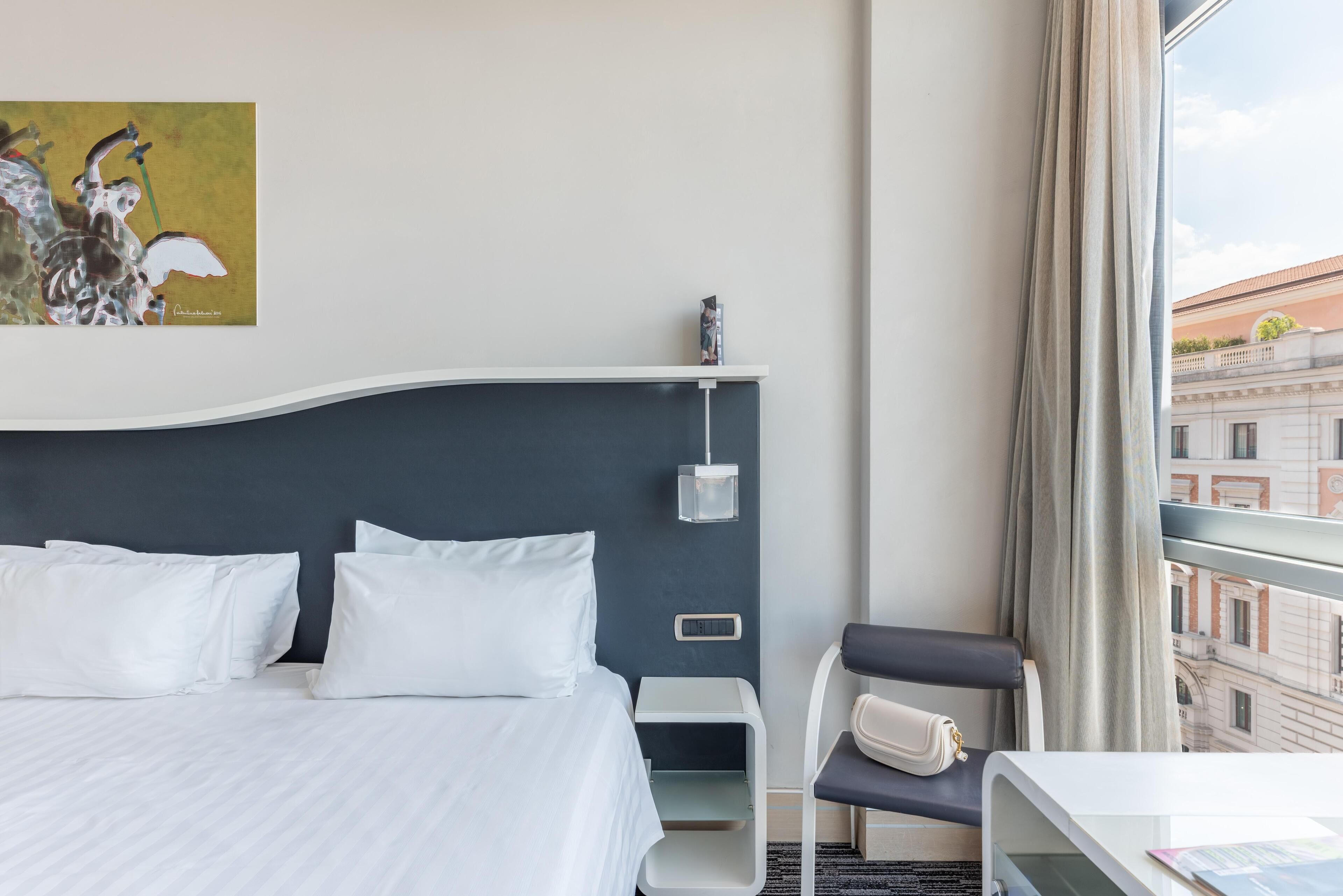 Una habitación de hotel con cama, una silla, un escritorio, un cuadro en la pared y vistas a edificios a través de la ventana.