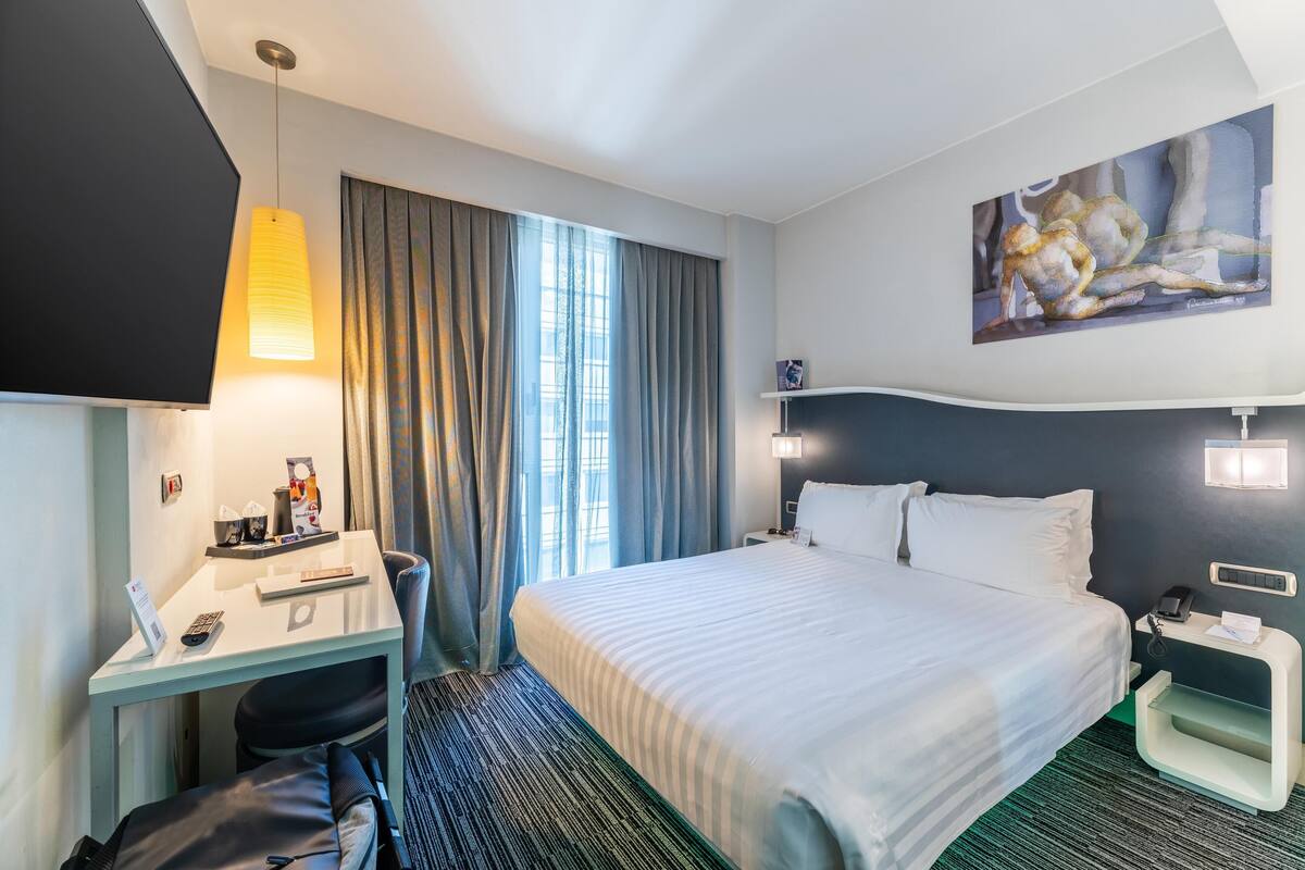 Een hotelkamer met een bed, een bureau, een stoel, een televisie, een schilderij en een raam met gordijnen.