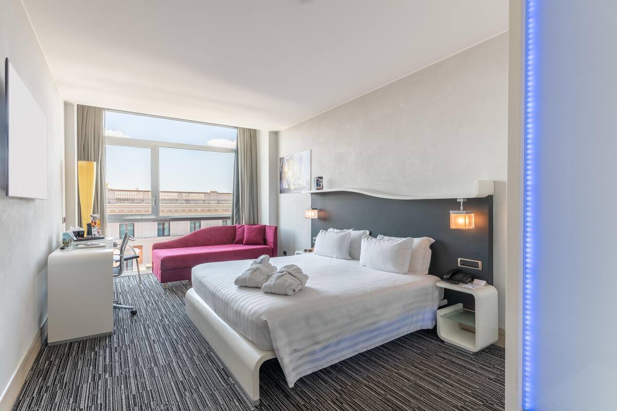 Een hotelkamer met een groot bed, een bureau, een stoel, een bank en een raam met uitzicht op gebouwen.