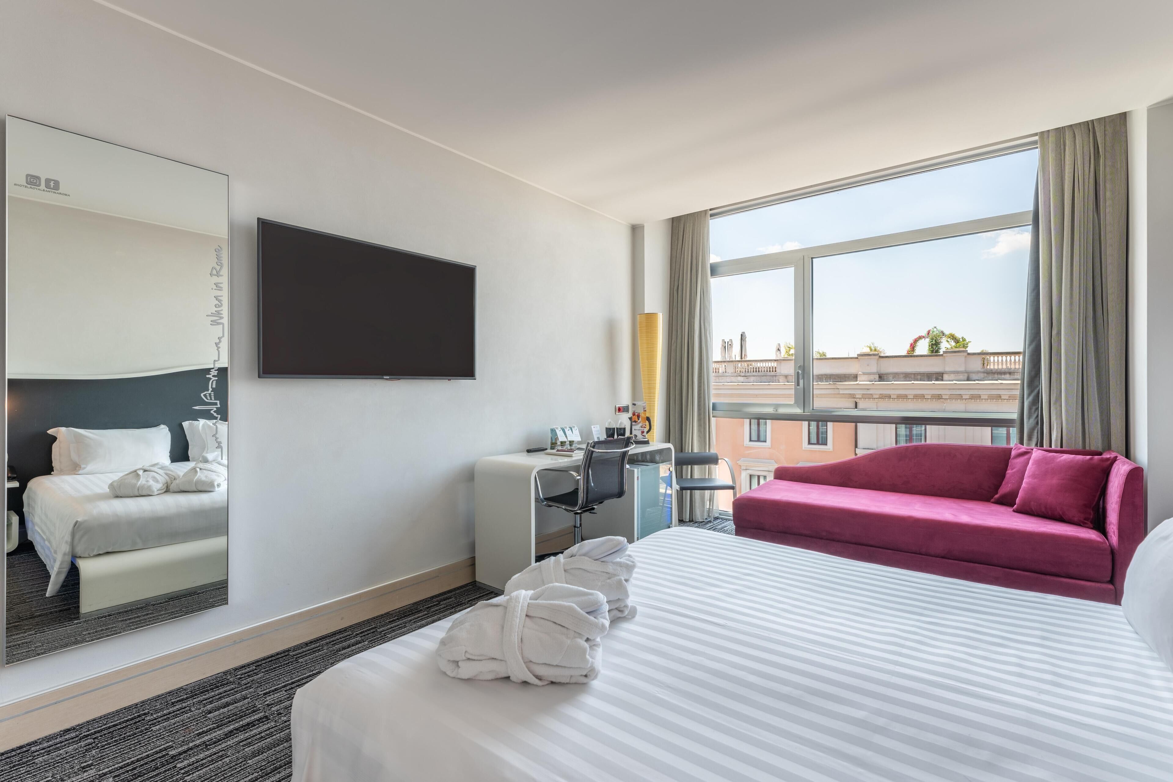 A hotel room with a bed, a desk, a chair, a sofa, a TV, and a large window with a city view.