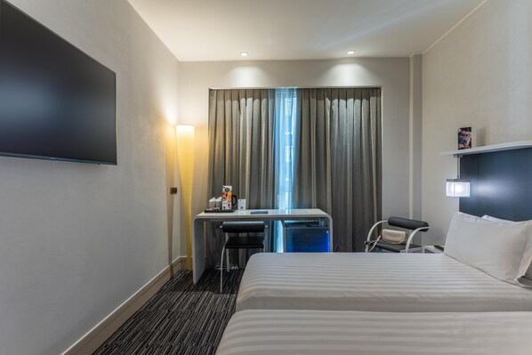 Een hotelkamer met een groot bed, een bureau met stoel, een flatscreen televisie, een klein tafeltje met lamp en een raam met gordijnen.
