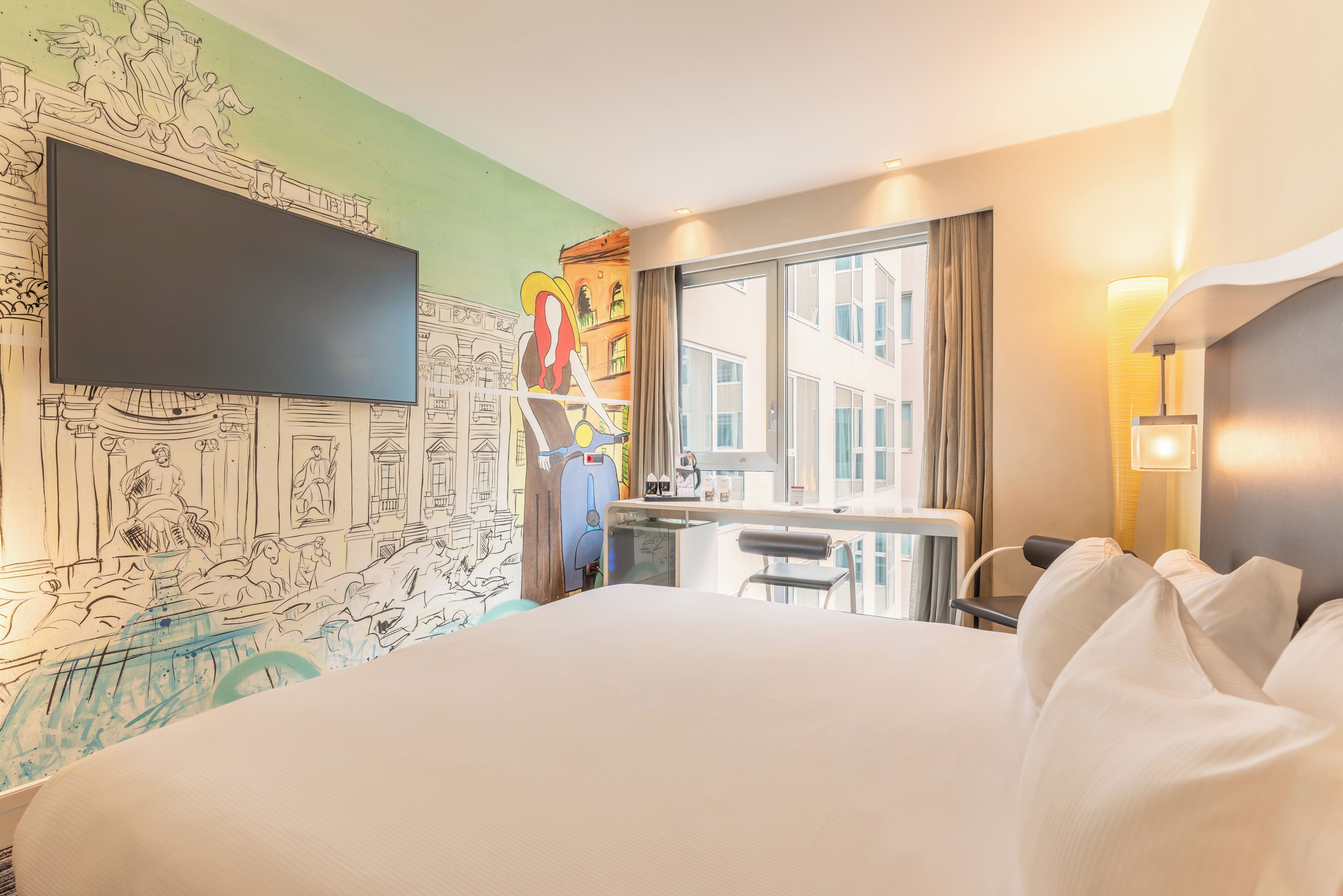 Camera d'albergo con un letto grande, una scrivania, una sedia, una televisione e una finestra con vista sugli edifici.