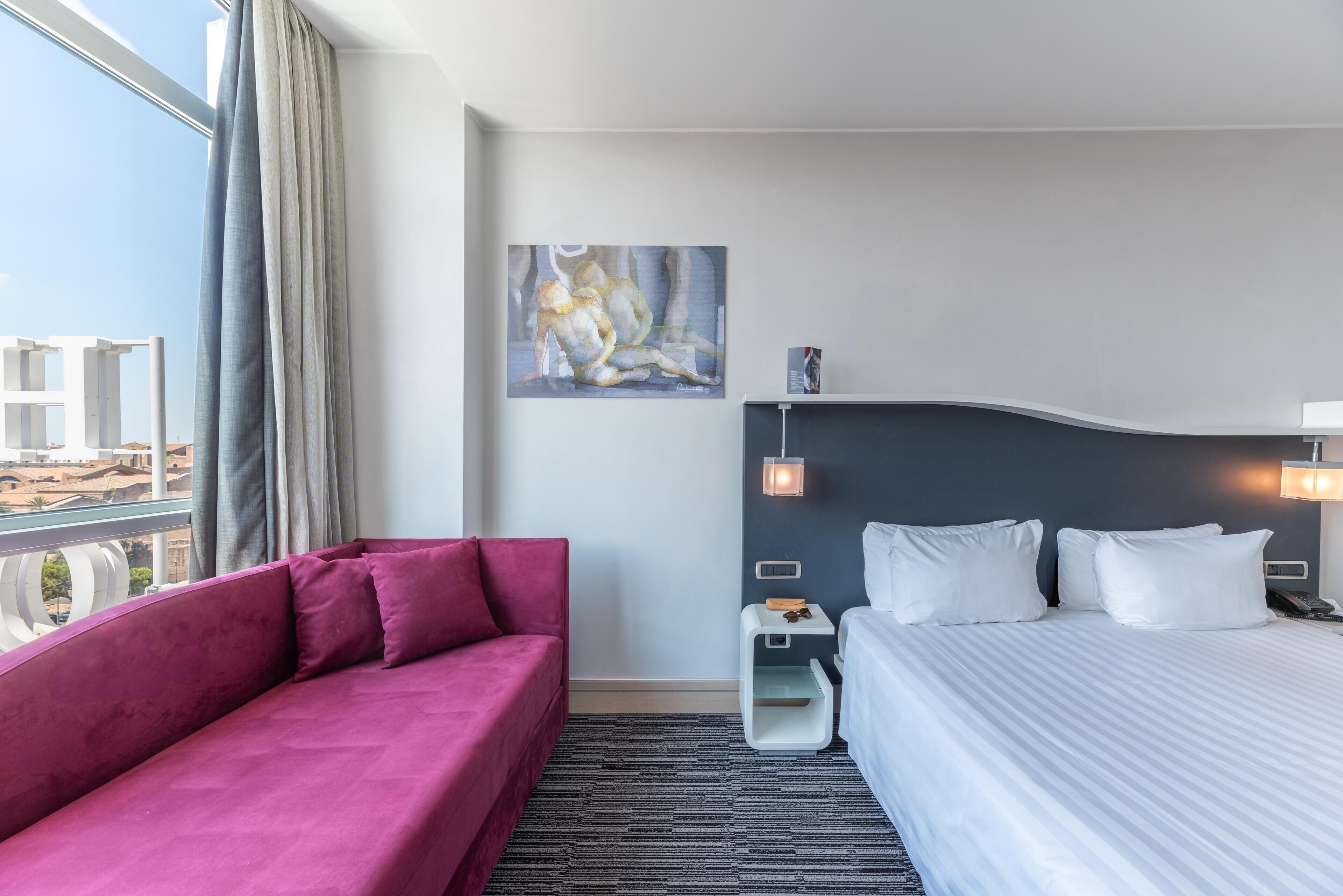 A modern hotel room with a large bed, a red sofa, a painting on the wall, and a city view.
