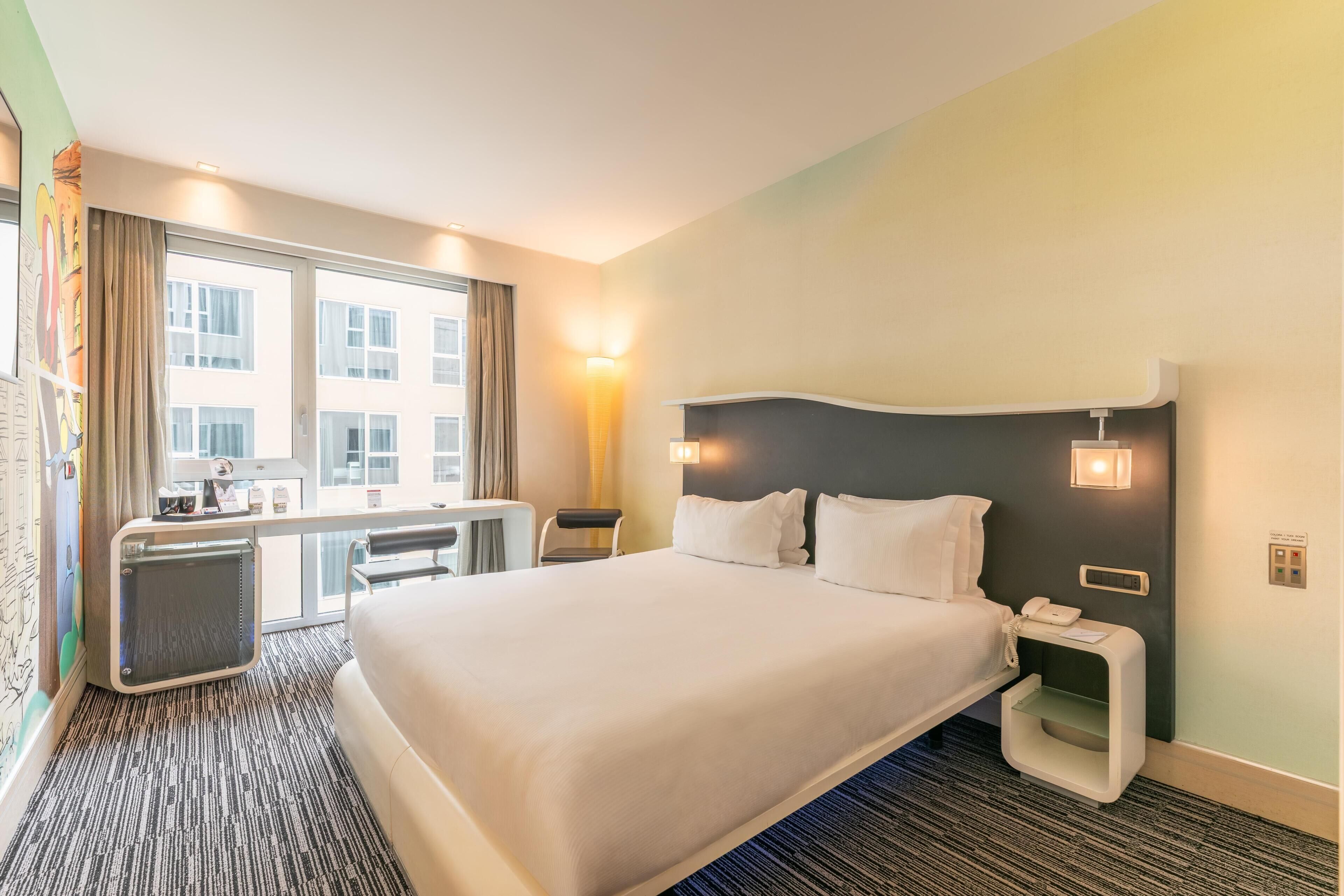 Ein Hotelzimmer mit einem großen Bett, einem Schreibtisch, einem Sessel, einem kleinen Tischchen, einem Nachttisch und einem Fenster mit Vorhängen.
