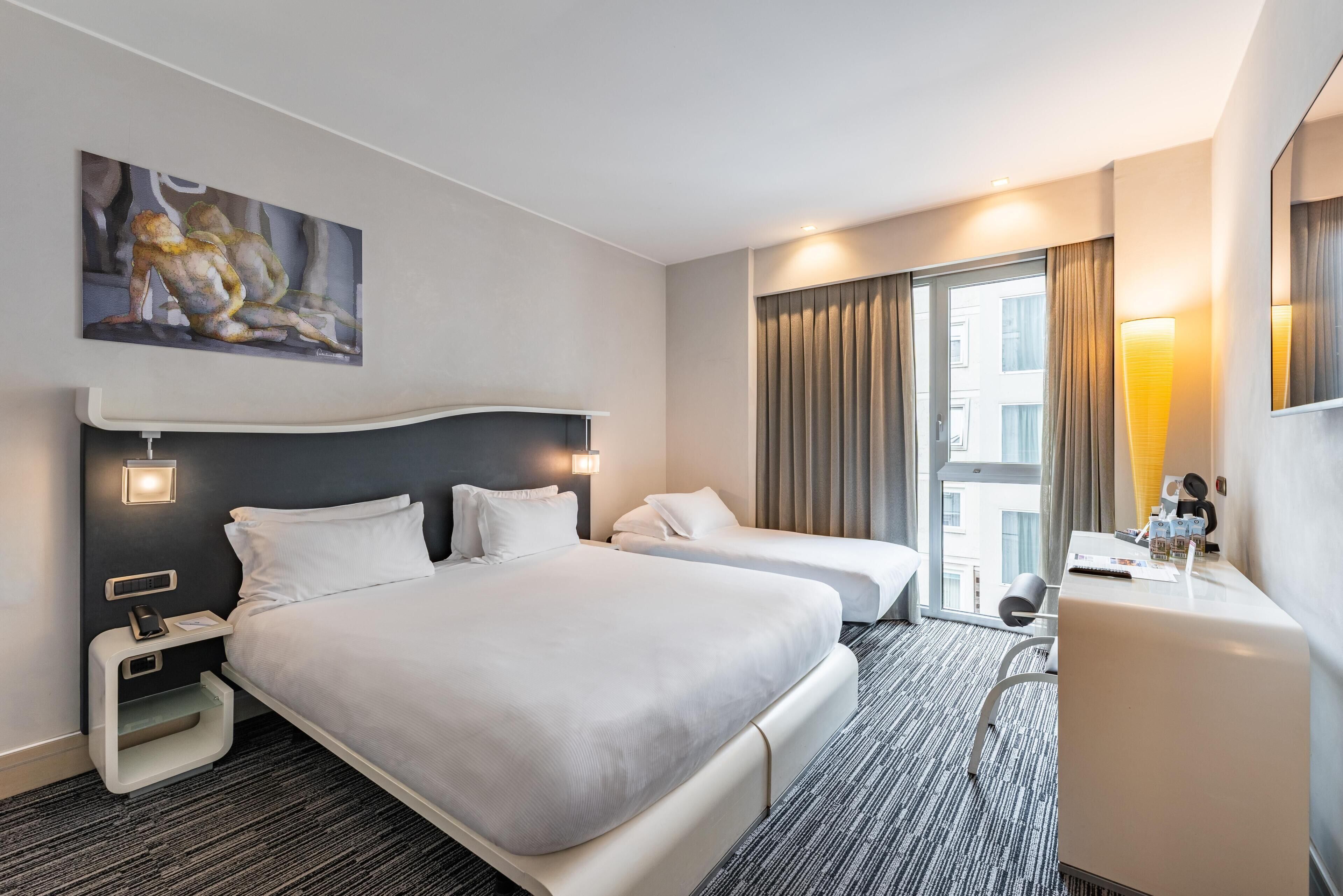 Habitación de hotel con una cama grande, dos mesitas de noche, un escritorio, una silla, un cuadro en la pared y una ventana con cortinas.