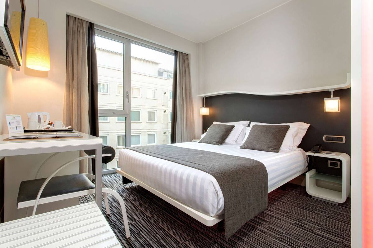 Hotelkamer met een bed, nachtkastje, lamp en uitzicht op gebouwen.