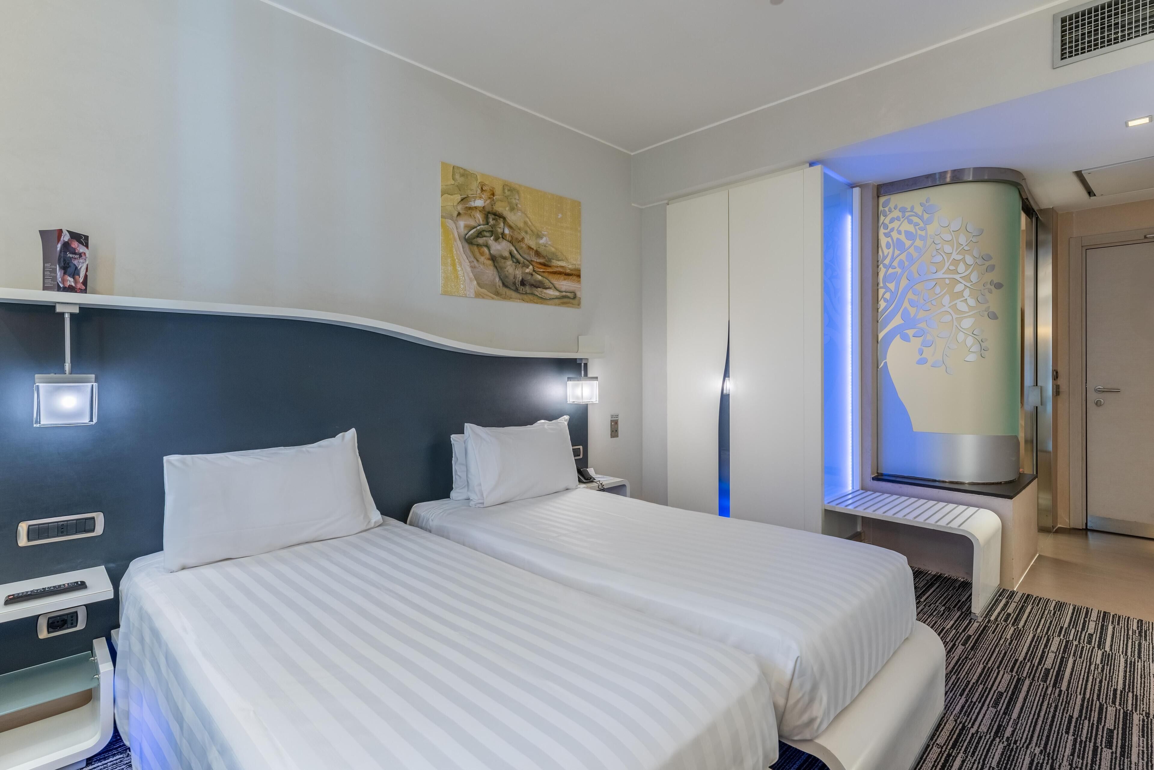 A hotel room with two beds, a large painting on the wall, a decorative wall panel, and a desk with a lamp.
