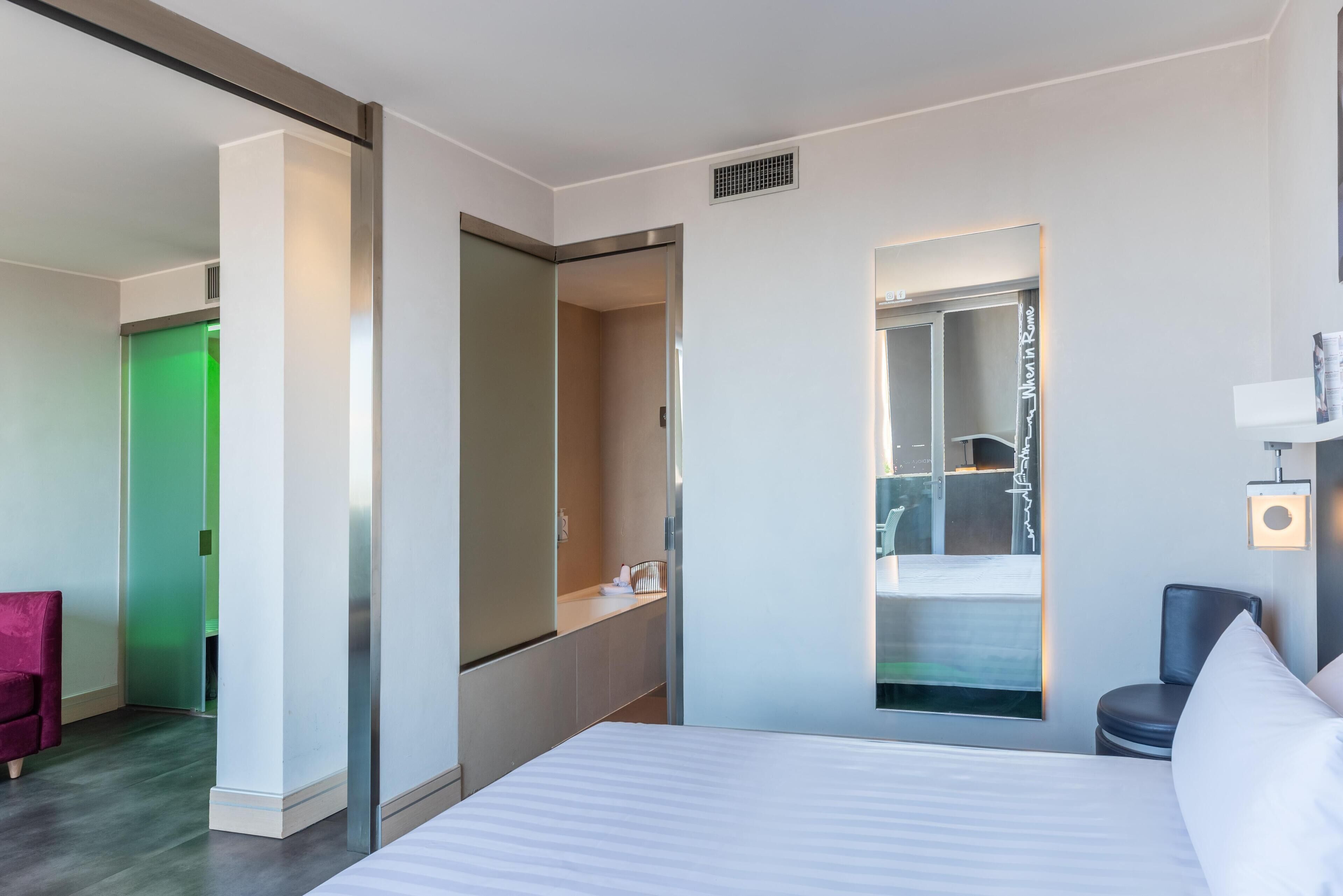 A modern hotel room with a large bed, a mirror, a chair, and a bathroom visible through a glass partition.