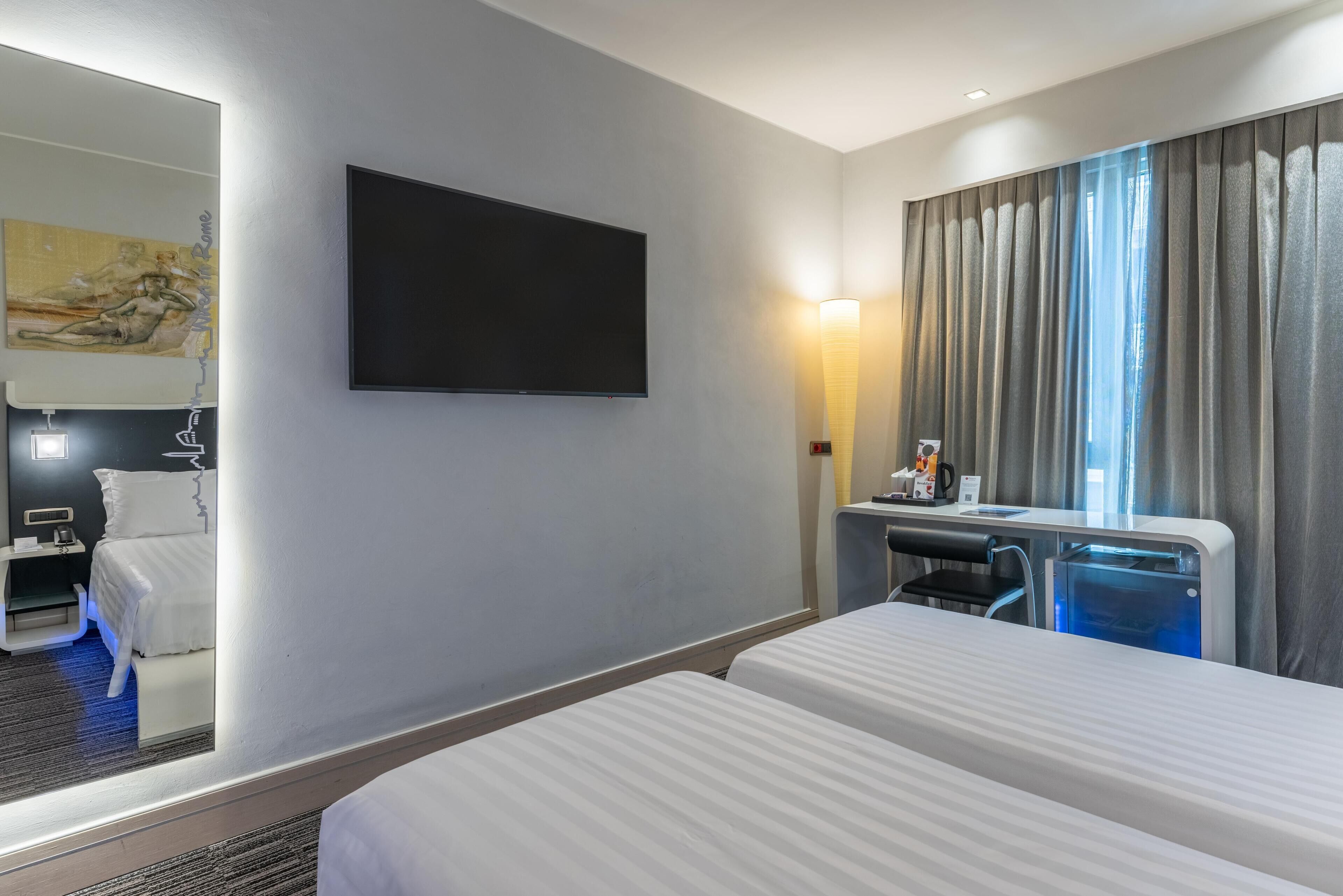 Une chambre d’hôtel avec deux lits, un téléviseur à écran plat, un bureau avec une lampe, un minibar et une grande fenêtre avec des rideaux.