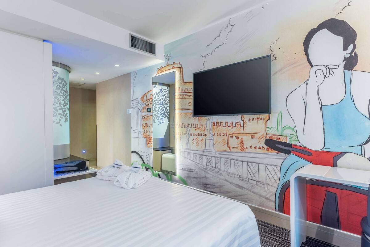 Een hotelkamer met een bed, een flatscreen televisie aan de muur, een groot muurschildering en een bureau met stoel.
