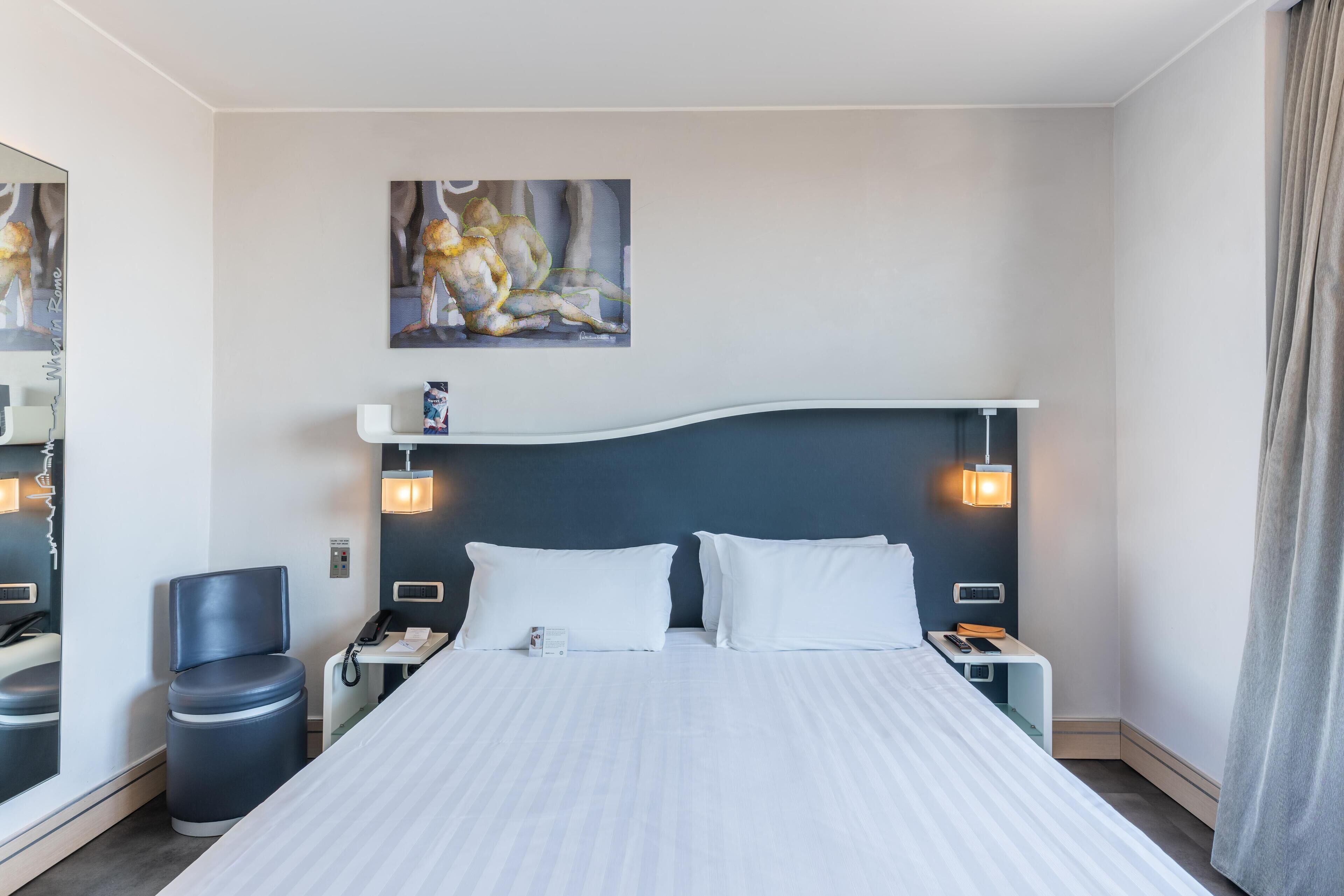 A hotel room with a large bed, two bedside tables, a nightstand, a chair, a trash bin, a painting on the wall, and a window with curtains.