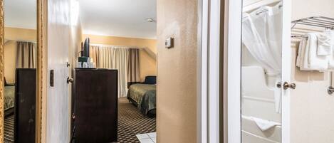 Quarto de hotel com cama, escrivaninha, espelho e banheiro com banheira e porta-toalhas.